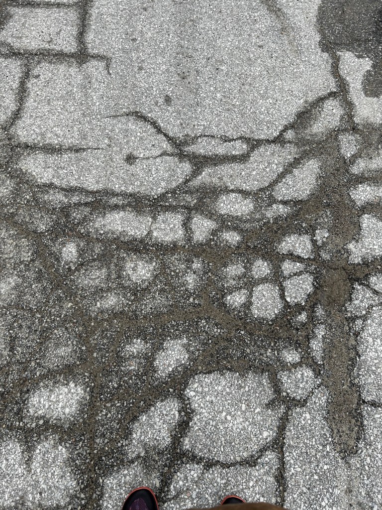 A road, cracked with wet areas, and the toes of shoes