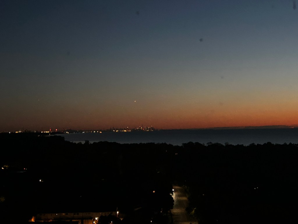 Nighttime just past sunset, lights visible on the horizon of the lake, trees with a few lights visible and a dark road.
